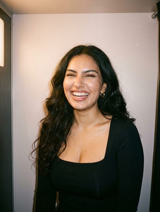 A smiling young woman with dark wavy hair wearing a black square-neck top in a photobooth